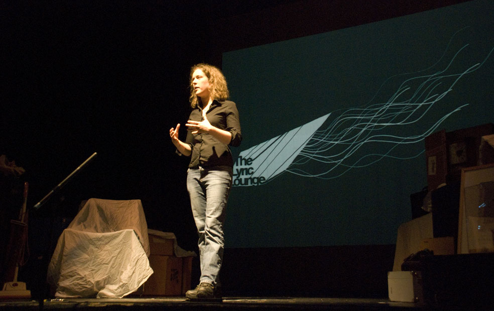 A slender, clean-shaven, white person with long, curly, dark blonde hair, wearing a black, fitted shirt with the sleeves rolled up, and faded blue jeans. They are standing, gesticulating, spotlit on a stage with a curved logo projected onto a dark screen behind them so that it looks as though they have a wing sprouting from their lower back. The logo reads The Lyric Lounge.