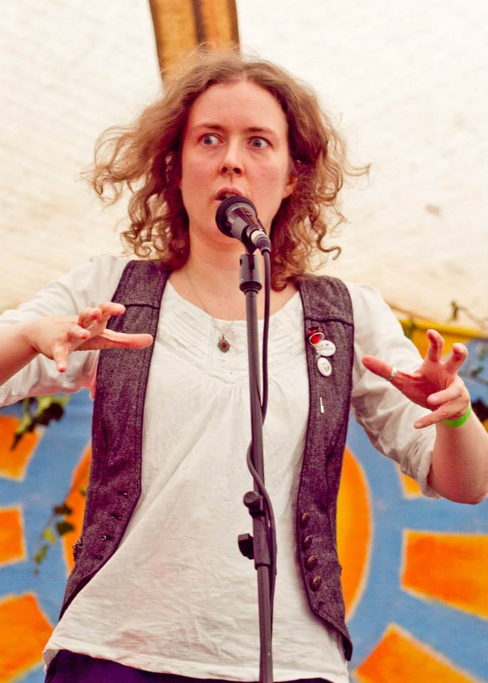 A white, clean-shaven person with slightly wild, curly, dark blonde hair is standing behind a mic on a stand, gesturing as though their hands were giant claws while looking askance. They're standing in a white tent in daylight, with a stylised orange sun in a blue sky on a banner decorated with ivy leaves behind them. They're wearing a white shirt with elbow-length sleeves, and a grey, tweed waistcoat with several badges on one lapel.