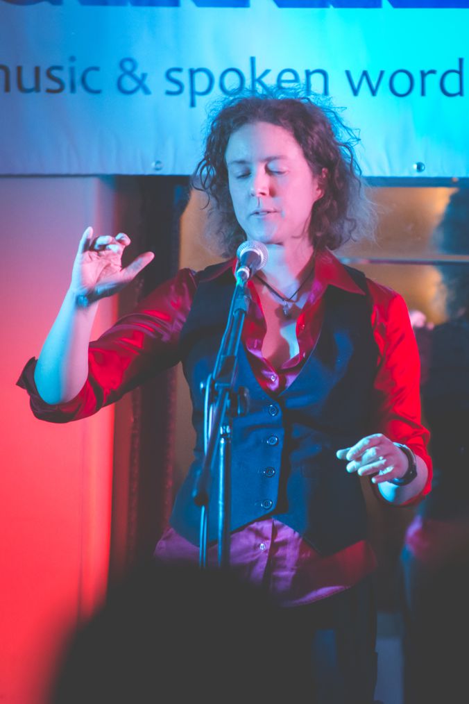 A vividly coloured photo of a white, clean-shaven person with collar-length, curly, light brown hair, gesturing with their eyes closed from behind a microphone on a stand. The are spotlit in pink and aquamarine, wearing a bright red, silky looking, fitted shirt under a formal, black waistcoat