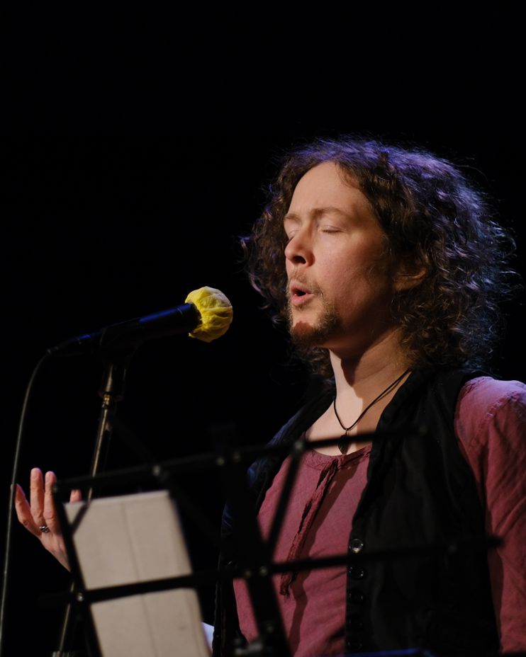 A white, bearded person with shoulder-length, curly, light brown hair, is standing spotlit on a dark stage behind a microphone on a stand; the mic has a yellow cover over it. The performer has their eyes lightly closed and their eyebrows raised slightly as if in slightly tortured contemplation. Their mouth is open as if they're saying "oh" or "ooh". From behind the basic music stand in front of them you can see one hand raised, palm-upward, as if cupping something. They are wearing a red shirt, black waistcoat, and a teardrop-shaped stone on a black cord around their neck. Their beard is a goatee, close-cropped.
