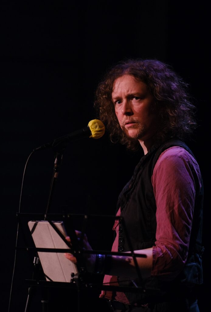 A white, bearded person with chin-length, curly hair of an indeterminate colour frowns suspiciously over their shoulder from behind a microphone stand. They are spotlight in a maroonish colour, wearing a dark red top and black waistcoat.