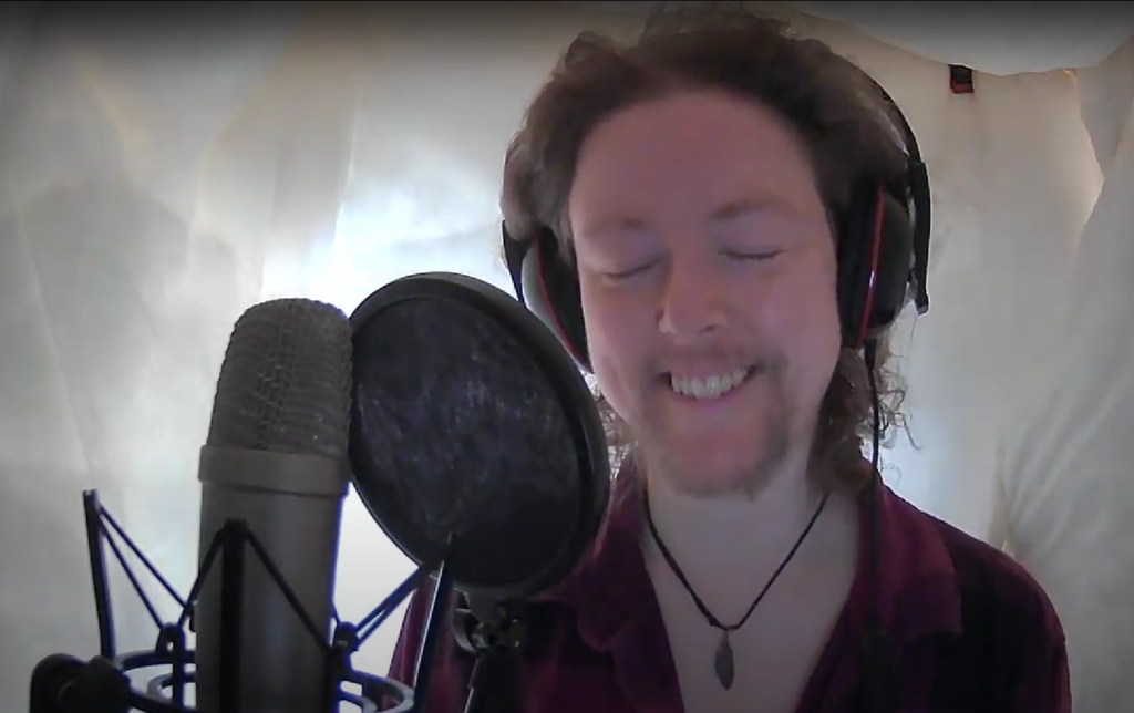 A webcam shot of a white, bearded person with light brown, collar-length, curly hair, smiling with their eyes closed from behind a pop-shielded vocal mic. They are wearing a dark red, plaid shirt, a tear-shaped, dark, stone pendant on a black cord around their neck, and large pair of black headphones with a red trim. Surrounding them are the white walls of a series of duvets, through which light is dimly shining.