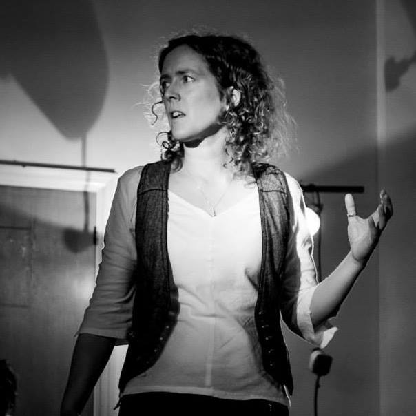 A black and white picture of Fay, a clean-shaven, white person with very curly, shoulder-length hair, performing indoors. They are strangely lit from several angles, with odd shadows thrown on the wall and door behind them. They are facing off to the left of the viewer, over their shoulder, with a somewhat anguished expression on their face, away from their raised, cupped hand. They are wearing a white, v-necked, linen shirt with capped sleeves, and an open, grey tweed waistcoat, a delicate metal pendant on an almost invisible chain, and a silver thumb ring on the upraised hand.